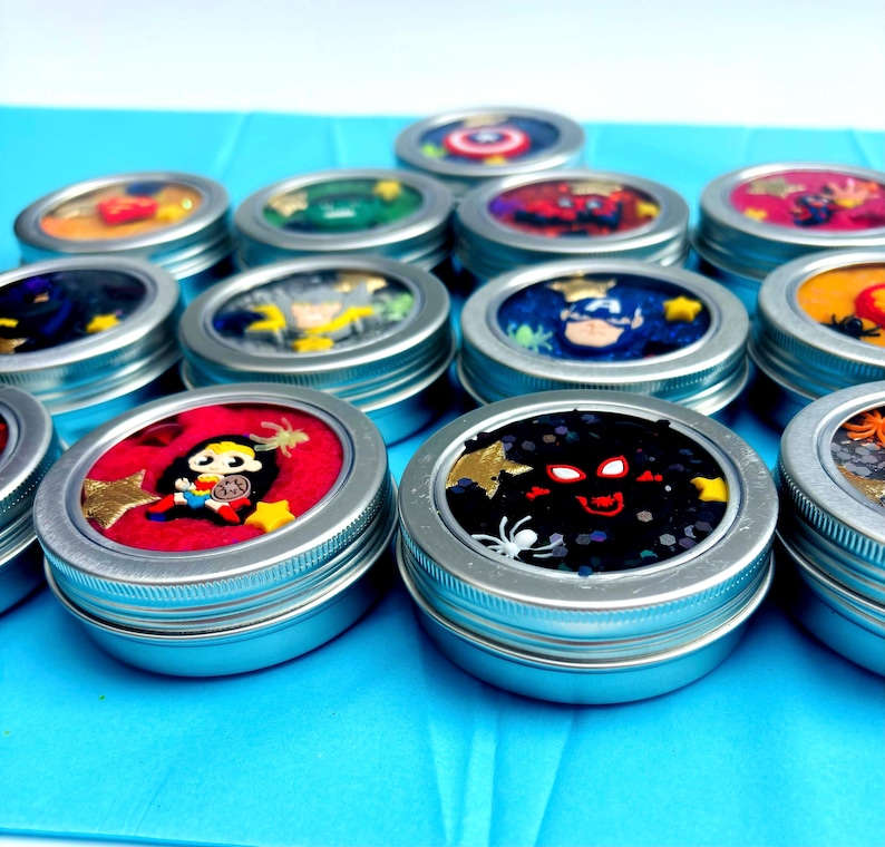 Puede incluir: Una colecci&oacute;n de peque&ntilde;os botes con tapas plateadas llenos de sustancias coloridas y brillantes. Cada bote presenta un dise&ntilde;o diferente con tem&aacute;tica de superh&eacute;roes. Los botes est&aacute;n dispuestos sobre una superficie azul brillante.