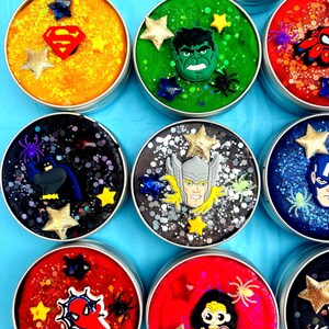 Puede incluir: Una colecci&oacute;n de slime colorido y brillante en peque&ntilde;os botes redondos de metal. Cada bote est&aacute; decorado con un emblema o personaje de superh&eacute;roe, como Superman, Hulk y Spider-Man, junto con adornos en forma de estrella.