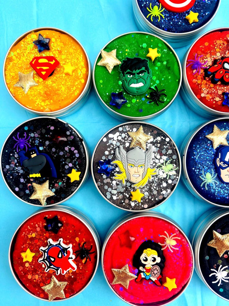 Puede incluir: Una colecci&oacute;n de slime colorido y brillante en peque&ntilde;os botes redondos de metal. Cada bote est&aacute; decorado con un emblema o personaje de superh&eacute;roe, como Superman, Hulk y Spider-Man, junto con adornos en forma de estrella.