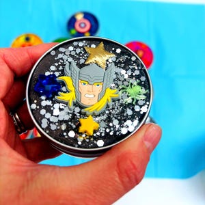 Puede incluir: Un recipiente redondo y negro lleno de purpurina y elementos decorativos. El recipiente presenta un adorno del personaje de Thor, una estrella dorada y una gema azul. Otros objetos coloridos son visibles en el fondo.