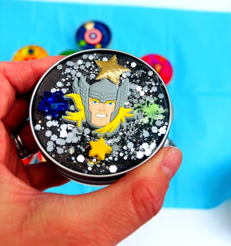 Puede incluir: Un recipiente redondo y negro lleno de purpurina y elementos decorativos. El recipiente presenta un adorno del personaje de Thor, una estrella dorada y una gema azul. Otros objetos coloridos son visibles en el fondo.