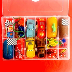 Może przedstawiać: Przezroczysty plastikowy organizer wypełniony różnymi zabawkami i akcesoriami. Zawiera zabawki samochodowe, zielonego węża, żółtego skorpiona i inne kolorowe przedmioty. Organizer stoi na czerwonej powierzchni.