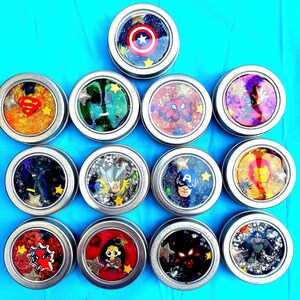 May include: Twelve small, round tins with clear lids, each filled with colourful, glittery substances and superhero designs. The designs feature American Hero’s shield, web crawler, and other superhero characters. The tins are arranged on a bright blue surface.