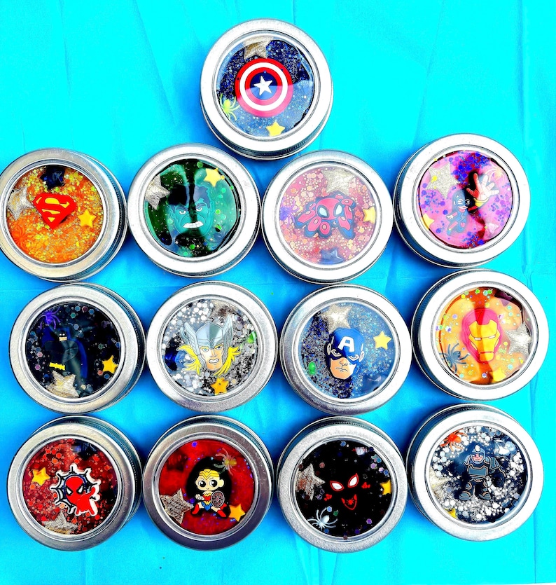 May include: Twelve small, round tins with clear lids, each filled with colourful, glittery substances and superhero designs. The designs feature American Hero’s shield, web crawler, and other superhero characters. The tins are arranged on a bright blue surface.