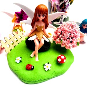 Peut inclure: Une figurine de fée aux cheveux roux et aux ailes blanches est assise sur une base verte avec une coccinelle, des fleurs et un champignon. Un buisson de fleurs roses et d'autres figurines de fées sont en arrière-plan. Un bocal avec des autocollants de fées est également visible.
