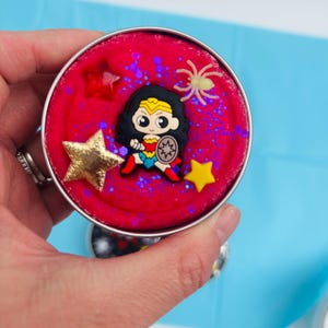 Puede incluir: Un recipiente redondo y plateado lleno de material rojo, decorado con una figura de Wonder Woman, adornos en forma de estrella y purpurina. Tambi&eacute;n hay una peque&ntilde;a ara&ntilde;a brillante. El recipiente se sostiene sobre un fondo azul.
