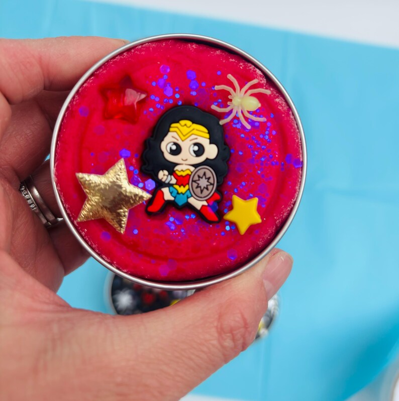 Puede incluir: Un recipiente redondo y plateado lleno de material rojo, decorado con una figura de Wonder Woman, adornos en forma de estrella y purpurina. Tambi&eacute;n hay una peque&ntilde;a ara&ntilde;a brillante. El recipiente se sostiene sobre un fondo azul.
