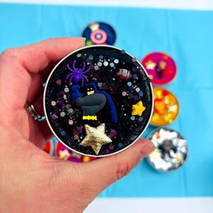 May include: A circular, black container featuring a Bat hero figure, a purple spider, and gold stars. The container is filled with glitter and decorative elements. Other colourful containers are in the background.