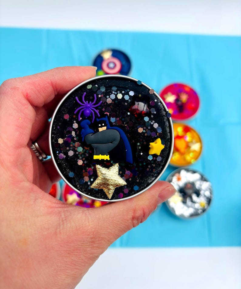 May include: A circular, black container featuring a Bat hero figure, a purple spider, and gold stars. The container is filled with glitter and decorative elements. Other colourful containers are in the background.