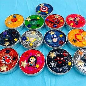 Puede incluir: Una colecci&oacute;n de slime colorido y brillante en peque&ntilde;os botes redondos de metal. Cada bote est&aacute; decorado con un personaje de superh&eacute;roe diferente, incluyendo el Capit&aacute;n Am&eacute;rica, Spider-Man y Wonder Woman, con estrellas y ara&ntilde;as.