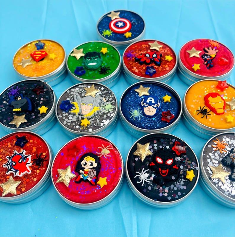 Puede incluir: Una colecci&oacute;n de slime colorido y brillante en peque&ntilde;os botes redondos de metal. Cada bote est&aacute; decorado con un personaje de superh&eacute;roe diferente, incluyendo el Capit&aacute;n Am&eacute;rica, Spider-Man y Wonder Woman, con estrellas y ara&ntilde;as.