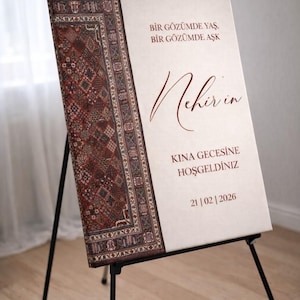 May include: A sign on a black easel with text in burgundy. The left side of the sign has a patterned rug design. The text reads "Bir Gözümde Yaş, Bir Gözümde Aşk", "Nehir'in", "Kına Gecesine Hoşgeldiniz", and "21|02|2026".
