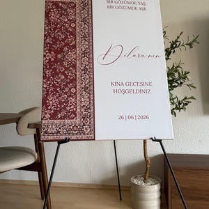May include: A large white sign on a black easel with text in Turkish and the date 26|06|2026. The left side of the sign features a burgundy and cream-colored patterned design.