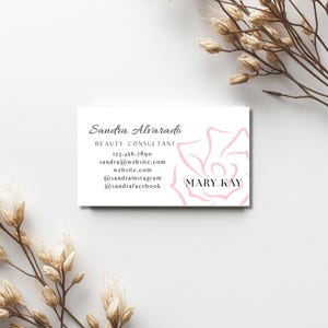 May include: A white business card with the name "Sandra Alvarado" in elegant script. The card includes contact information and a pink floral design with the "MARY KAY" logo. Dried floral stems frame the card on a white background.