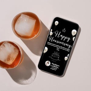Könnte beinhalten: Zwei Gläser mit Eiswürfeln und bernsteinfarbener Flüssigkeit liegen neben einem Smartphone. Das Telefon zeigt einen schwarzen Bildschirm mit weißem Text "Happy Housiversary!" und zeigt einen Kuchen und Luftballons.
