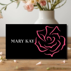 May include: A black business card featuring the white text "MARY KAY" and a pink rose outline. The card rests on a wooden surface, with a white vase and flowers in the background.