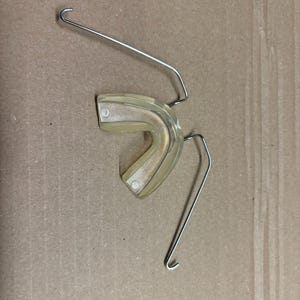 Puede incluir: Un retenedor dental claro y curvo con dos extensiones de alambre metálico. El retenedor está hecho de un material translúcido y está diseñado para encajar sobre los dientes. Los alambres metálicos tienen extremos en forma de gancho.