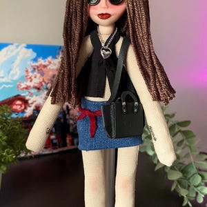 Puede incluir: Mu&ntilde;eca hecha a mano con ojos de bot&oacute;n, cabello casta&ntilde;o y l&aacute;piz labial rojo. La mu&ntilde;eca lleva un chaleco negro, una falda vaquera y un peque&ntilde;o bolso negro. Un colgante de coraz&oacute;n plateado cuelga del cuello de la mu&ntilde;eca. Los zapatos de la mu&ntilde;eca son negros.