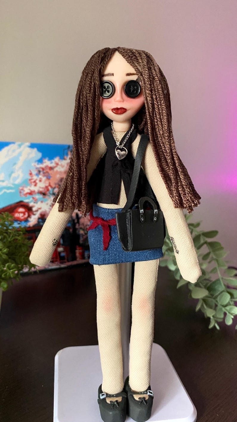 Puede incluir: Mu&ntilde;eca hecha a mano con ojos de bot&oacute;n, cabello casta&ntilde;o y l&aacute;piz labial rojo. La mu&ntilde;eca lleva un chaleco negro, una falda vaquera y un peque&ntilde;o bolso negro. Un colgante de coraz&oacute;n plateado cuelga del cuello de la mu&ntilde;eca. Los zapatos de la mu&ntilde;eca son negros.