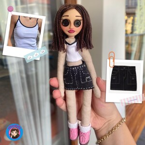 Puede incluir: Mu&ntilde;eca hecha a mano con ojos de bot&oacute;n, que lleva un top corto blanco con ribete negro y una falda vaquera negra con costuras blancas. La mu&ntilde;eca tiene pelo de hilo marr&oacute;n y zapatos rosas. Una peque&ntilde;a foto de una camiseta sin mangas blanca est&aacute; en el fondo.