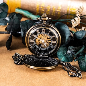 May include: A bronze-toned mechanical pocket watch with a black face featuring white Roman numerals and an exposed gold-colored gear movement. The watch has a circular case, a winding crown at the top, and an attached metal chain link.