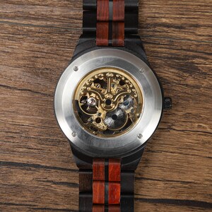 Op de afbeelding: Een houten polshorloge met een zwart frame en een bruine houten band. De wijzerplaat is zichtbaar en toont het ingewikkelde mechanische uurwerk met goudkleurige tandwielen. Het horloge wordt gepresenteerd op een houten oppervlak.