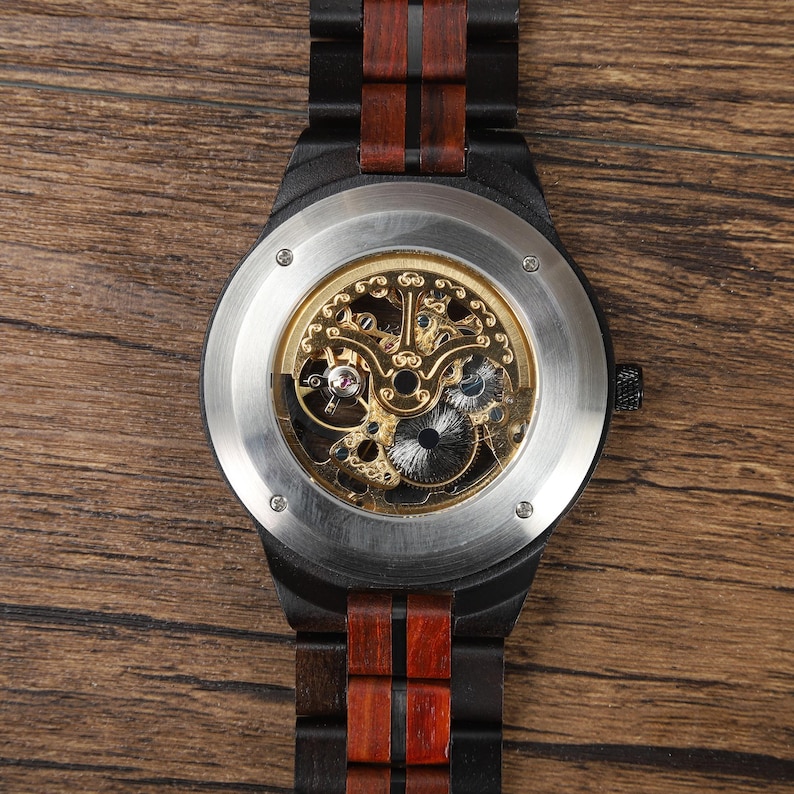 Op de afbeelding: Een houten polshorloge met een zwart frame en een bruine houten band. De wijzerplaat is zichtbaar en toont het ingewikkelde mechanische uurwerk met goudkleurige tandwielen. Het horloge wordt gepresenteerd op een houten oppervlak.