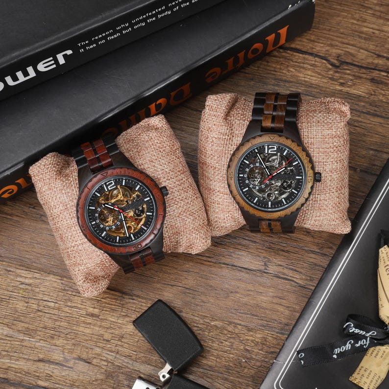 Op de afbeelding: Twee houten polshorloges met zichtbare binnenwerken worden gepresenteerd op kleine burlap kussens. De horloges hebben donker hout en zwarte wijzerplaten, met het nummer 12 duidelijk zichtbaar. Ze liggen op een houten oppervlak.