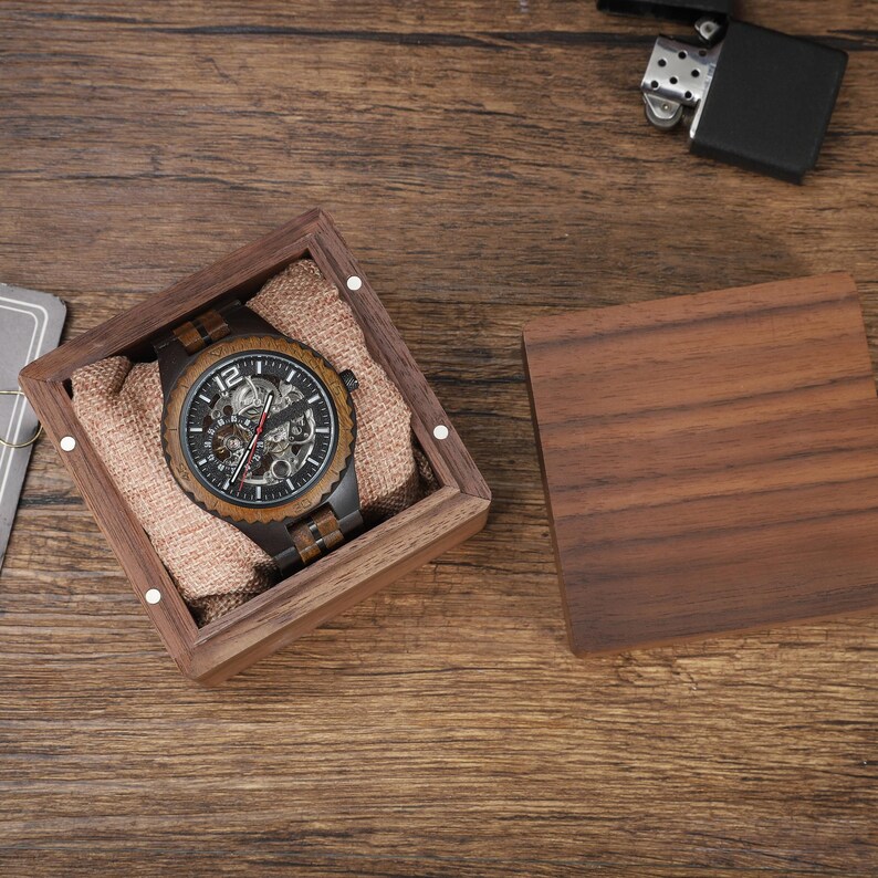 Op de afbeelding: Een houten horloge met een zwarte wijzerplaat en zichtbare mechanismen, gepresenteerd in een vierkante houten doos met een jute voering. Het horloge heeft een houten rand en een metalen band. De doos is open.