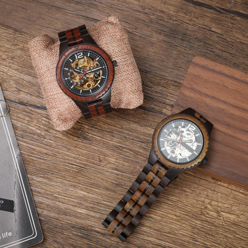 Op de afbeelding: Twee houten polshorloges met zichtbare tandwielen en donkere wijzerplaten. E&eacute;n horloge wordt weergegeven op een tan stoffen kussen. De horlogebanden en lunetten zijn gemaakt van donker hout met roodbruine accenten. De horloges liggen op een houten oppervlak.