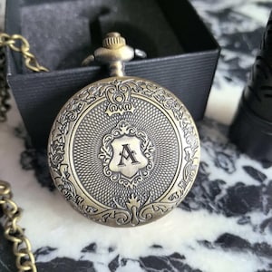 Könnte beinhalten: Antike Taschenuhr aus Messing mit einem verzierten Design. Die Uhr hat ein Schild mit dem Buchstaben "A" in der Mitte, umgeben von dekorativen Schnörkeln. Die Uhr ist an einer Kette und wird in einer schwarzen Box präsentiert.