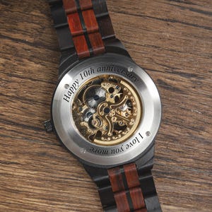 Op de afbeelding: Zwarte houten horloge met zichtbaar mechanisch uurwerk en roestvrijstalen achterkant. De horlogeband heeft donkere hout- en roodbruine accenten. Tekst op de achterkant luidt "Happy 10th anniversary" en "I love you more."