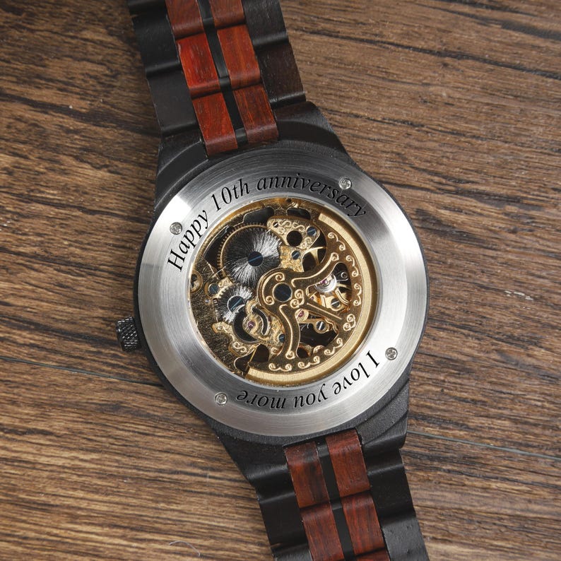 Op de afbeelding: Zwarte houten horloge met zichtbaar mechanisch uurwerk en roestvrijstalen achterkant. De horlogeband heeft donkere hout- en roodbruine accenten. Tekst op de achterkant luidt "Happy 10th anniversary" en "I love you more."