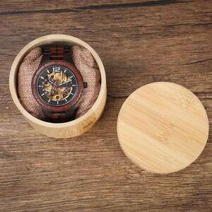 Op de afbeelding: Een houten polshorloge met een donkere wijzerplaat en zichtbare interne mechanismen ligt in een ronde bamboe doos met een stoffen voering. Het horloge heeft een bruine houten band en een zwarte bezel. De deksel ligt ernaast.