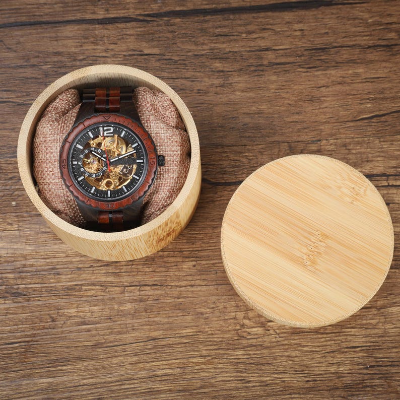 Op de afbeelding: Een houten polshorloge met een donkere wijzerplaat en zichtbare interne mechanismen ligt in een ronde bamboe doos met een stoffen voering. Het horloge heeft een bruine houten band en een zwarte bezel. De deksel ligt ernaast.