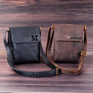 May include: Two leather crossbody bags. One is black with the name "Martin" and the letter "M" printed on the front. The other is brown with the name "J.Barlow" printed on the front. Both bags have adjustable straps and gold-colored hardware.