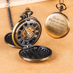 Peut inclure: Une montre de poche mécanique couleur bronze avec un cadran noir, des chiffres romains et un mécanisme apparent. Un couvercle assorti est suspendu au-dessus, gravé du texte : To my son: Whatever comes, I'll love you, just as I do now and forever.