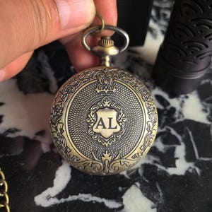 Pode incluir: Relógio de bolso antigo em latão com um design floral e de rolagem ornamentado. O mostrador do relógio apresenta um escudo com as iniciais "AL". O relógio tem formato circular, com uma haste de enrolamento e um pequeno laço para uma corrente.