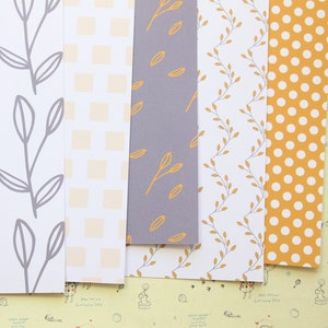 May include: Five sheets of patterned paper with different designs. The first sheet has a grey leaf pattern on a white background. The second sheet has a white and yellow chequered pattern. The third sheet has a grey background with a yellow leaf pattern. The fourth sheet has a white background with a yellow leaf pattern. The fifth sheet has a yellow background with white polka dots.