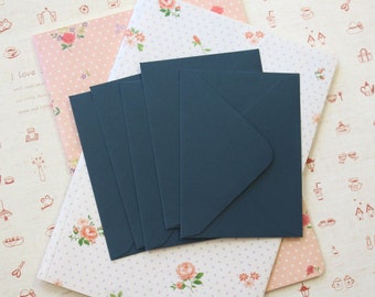 May include: A stack of five dark teal envelopes rests on a collection of patterned stationery. The stationery includes a pink notebook with a floral design and sheets with a repeating pattern of small flowers and dots. The background features a cream-colored sheet with red illustrations and the text "I love zakka".