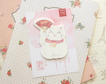 May include: A white lucky cat figurine with a red fan and a red collar. The cat is holding a red ball in its paw. The figurine is on a pink and white background with the text "Miss Time" in black.