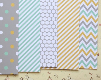 May include: Five sheets of patterned paper with different designs. The first sheet has a gray background with white and yellow polka dots. The second sheet has a light blue and white striped pattern. The third sheet has a gray background with white polka dots. The fourth sheet has a yellow background with white stripes. The fifth sheet has a gray and light blue chevron pattern.