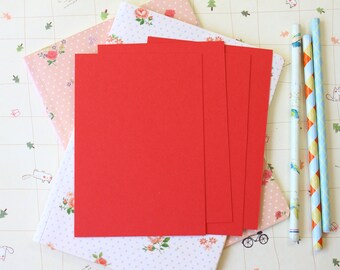May include: A stack of five red cardstock paper sheets on top of a floral patterned notebook. The notebook has white polka dots and pink flowers. Three striped straws are on the right side of the image.