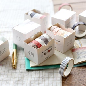May include: A set of five washi tape rolls in white boxes with the text "芯陌 SIMNO" printed on the front. The washi tape rolls are decorated with various patterns, including stripes, polka dots, and geometric designs. The boxes are arranged on a wooden surface with a notebook and a pen.