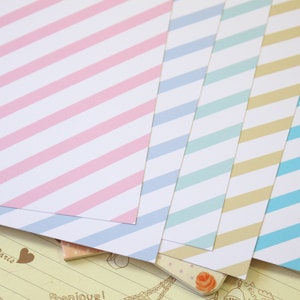 May include: Five sheets of decorative paper with diagonal stripes in pink, light blue, turquoise, beige, and blue. The paper is on a background of a page with a light brown grid pattern and the word "Paris" written in brown.