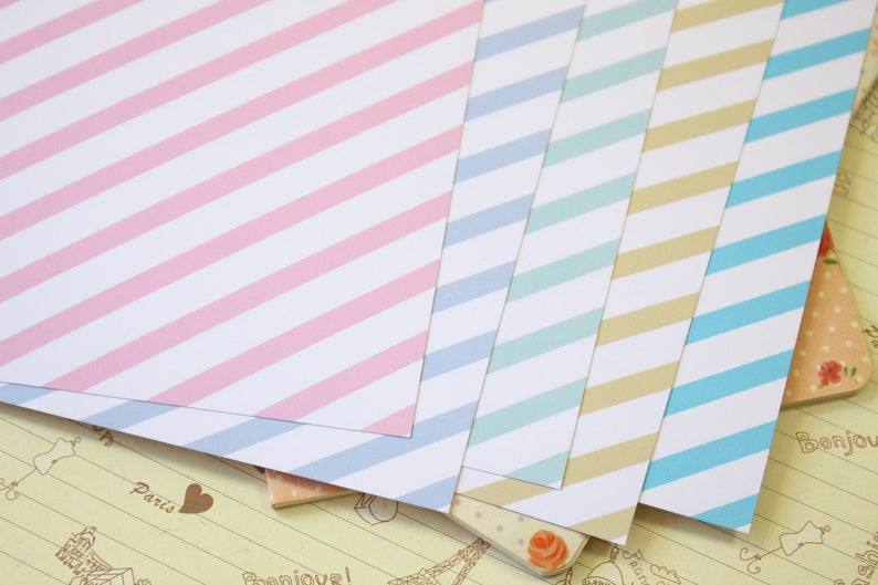 May include: Five sheets of decorative paper with diagonal stripes in pink, light blue, turquoise, beige, and blue. The paper is on a background of a page with a light brown grid pattern and the word "Paris" written in brown.
