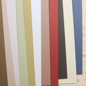 Puede incluir: Una colecci&oacute;n de 10 hojas de papel de diferentes colores, cada una con una textura &uacute;nica. Las hojas de papel est&aacute;n dispuestas en una pila vertical, con la hoja superior de color marr&oacute;n claro y la hoja inferior de color azul oscuro.