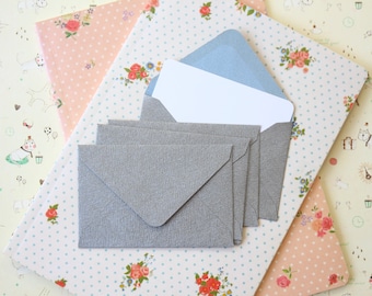 May include: A set of five gray envelopes with a textured finish, on a white background with blue polka dots and floral patterns. The envelopes are stacked on top of each other, with the top envelope slightly open to reveal a blank white card inside a light blue envelope.