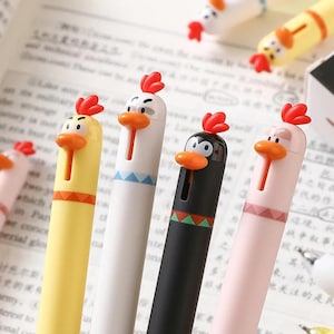 May include: A selection of novelty pens designed as cartoon chickens. The pens are in yellow, white, black, and pink, each with a red comb and orange beak. The pens have a slot for the pen tip, and are designed to be fun stationery items.