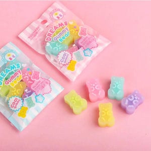 May include: Colorful gummy bear candies in various pastel colors, including pink, green, yellow, and purple, are scattered on a pink surface. Two clear plastic bags filled with gummy bears are also present, with text that reads "DREAMS Gummy Bear".